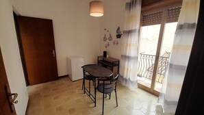 Una habitación pequeña con una puerta de madera, un refrigerador blanco, una mesa con dos sillas y una ventana con vistas al balcón.