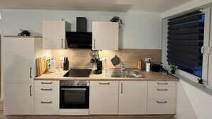 Une cuisine de taille réduite avec des meubles blancs, un four noir, un évier et une fenêtre équipée de stores.