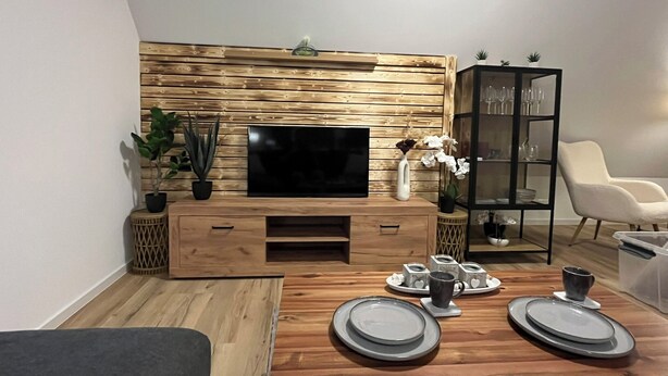 Un salon moderne avec un panneau mural en bois, un téléviseur à écran plat, une table à manger avec des assiettes et une tasse à café, et une chaise blanche.
