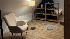 Une chambre à coucher confortable aménagée en mezzanine, avec un plafond en pente, un fauteuil moelleux, une lampe sur pied, une bibliothèque en bois et une petite plante.