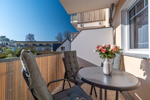 A balcony with a round table and two chairs, a vase with flowers, and a view of neighboring buildings.
