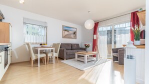 Ein modernes Wohnzimmer mit Sofa, Esstisch und Stühlen, einem Küchenbereich mit Herd und einem großen Fenster mit Vorhängen.