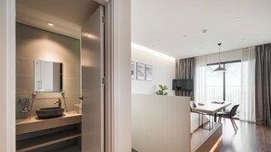 A modern hotel room with a wooden floor, a large window with curtains, a dining area with a table and chairs, and a bathroom with a large mirror and a vessel sink. - HN ETH Vinhomes D'capitale Resident (Hanoi)