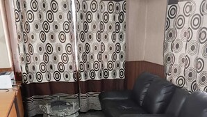A living room with a black leather sofa, a glass coffee table, and patterned curtains. - Hotel New Green view-apart (Srinagar)