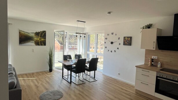 A modern kitchen with a dining area, wooden flooring, and a large window providing natural light. - FeWo Foorden Wohnglück 3 (Hesel)