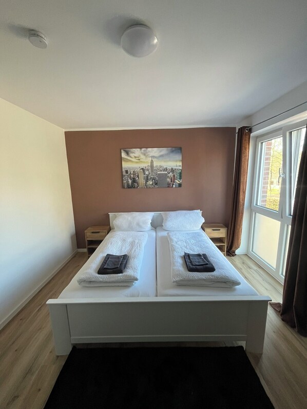 A double bed with white linens and towels, a cityscape painting on the wall, and a window with curtains.