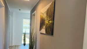 A hallway with wooden flooring, a potted plant, a rug, a painting of a landscape, and a geometric light fixture. - FeWo Foorden Wohnglück 3 (Hesel)