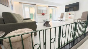 Un soggiorno moderno con divano, poggiapiedi e tavolo da pranzo. C’è una TV montata a parete e una ringhiera da balcone.
