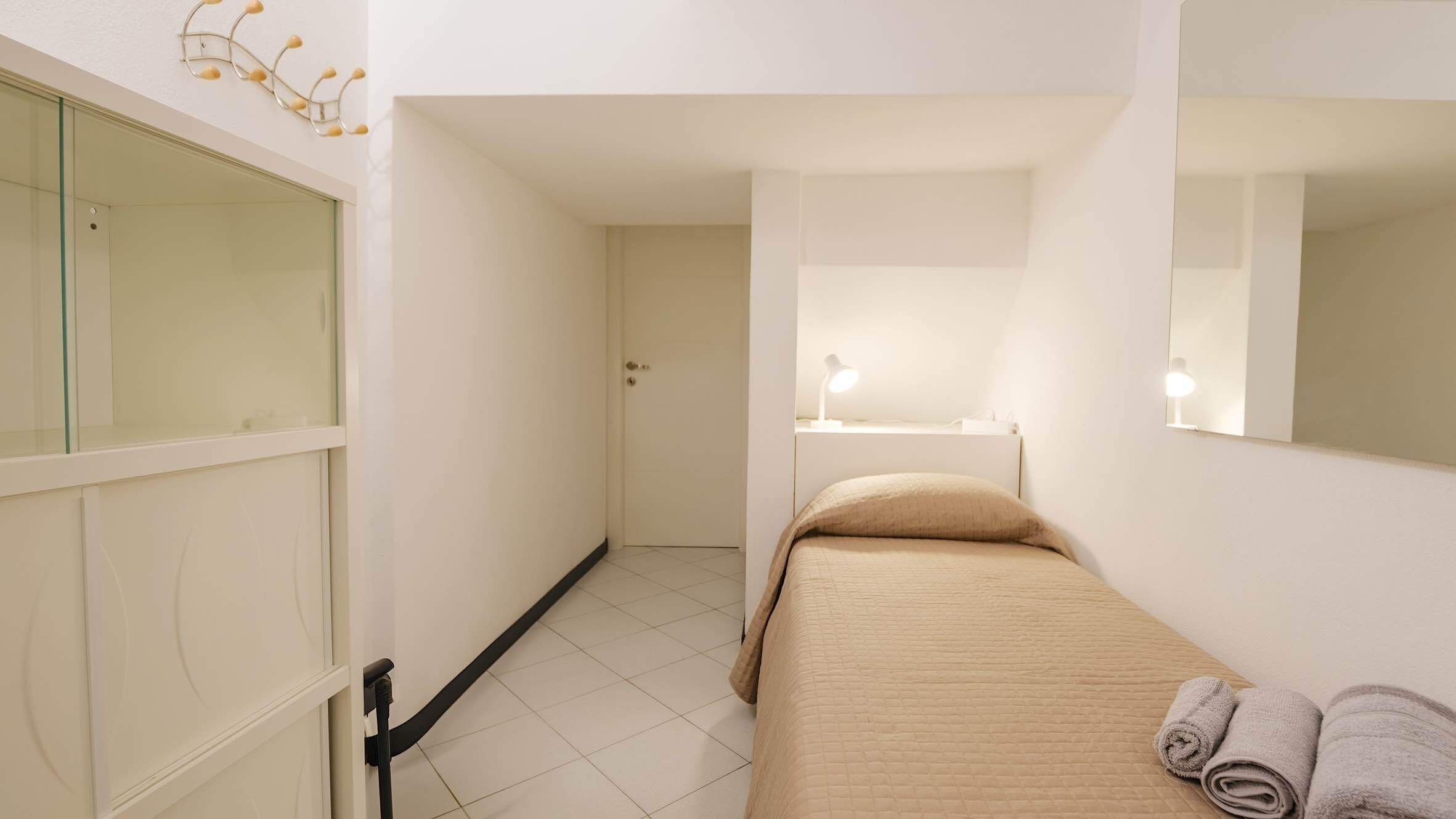 Une chambre d’hôtel étroite, avec un lit, une table de chevet sur laquelle se trouve une lampe, un miroir et une serviette posée sur le lit.