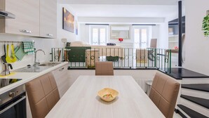 Una cucina moderna con zona pranzo, caratterizzata da un tavolo dai toni chiari, sedie imbottite marroni e vista sull'area soggiorno tramite una porta in vetro.