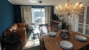 Een eetgedeelte met een houten tafel voor vier personen, omringd door stoelen. Boven de tafel hangt een kroonluchter, en er is een bank en fauteuil zichtbaar op de achtergrond.