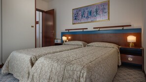 A hotel room with two beds, a wooden headboard, bedside lamps, a painting on the wall, and a door.