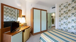 A hotel room with a bed, a desk with a lamp, a television, and a wardrobe.