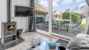 Una habitación acogedora con chimenea de leña, televisión y un amplio ventanal corredera que da a un patio exterior con mesa y sillas.