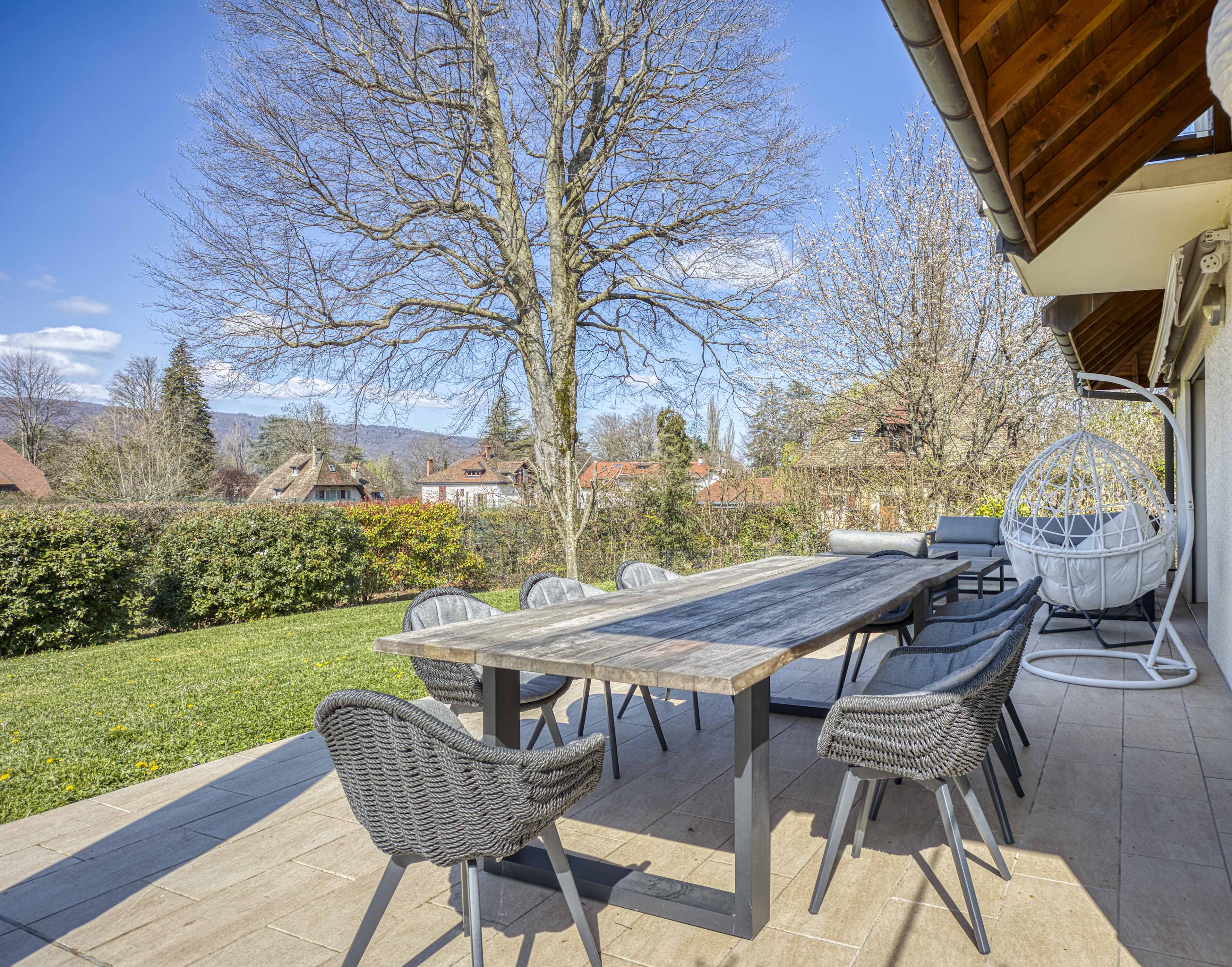 Een terras buiten met een houten tafel en rieten stoelen, een hangstoel, en in de achtergrond huizen en bomen.