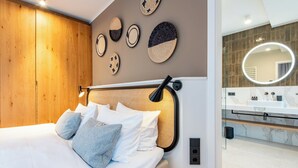Una camera da letto moderna con testiera in legno, un letto con lenzuola bianche e cuscini grigi, una parete decorata con opere d'arte in tessuto e un bagno con lavabo in marmo e specchio tondo.