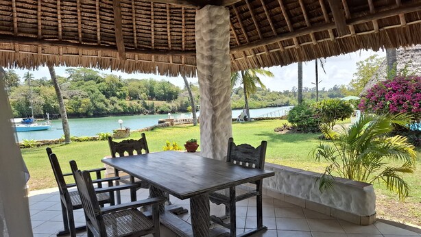 A covered outdoor seating area with a table and chairs, overlooking a lake and lush greenery. - Villa Dolphin (Mtwapa)