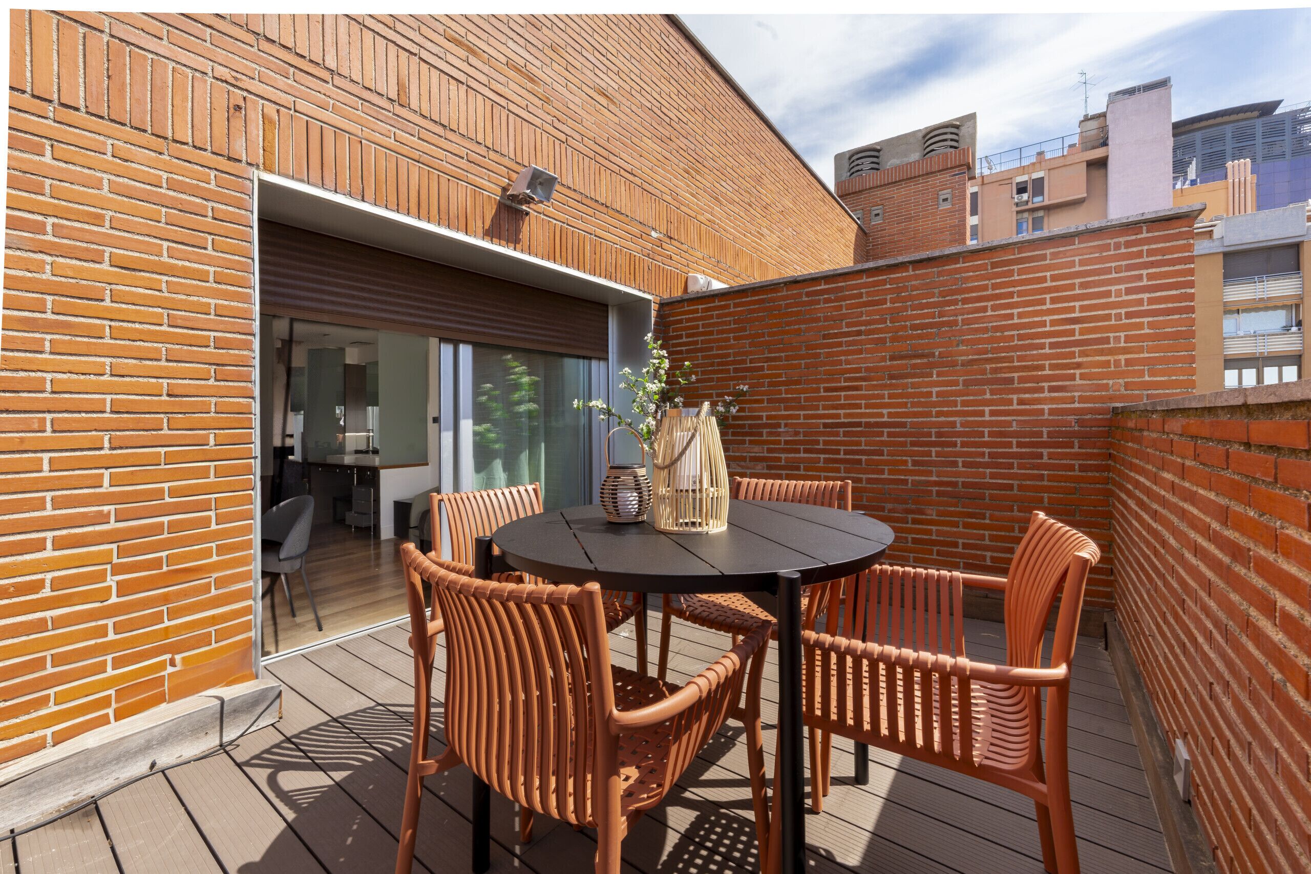 Une terrasse avec une table ronde et des chaises en osier, un mur en briques et une porte vitrée donnant sur un espace intérieur.