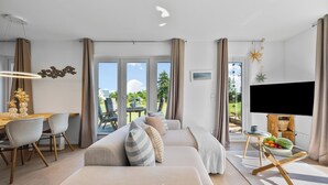 En moderne stue med sofa, spiseområde med stole, fjernsyn og en balkon med udsigt.