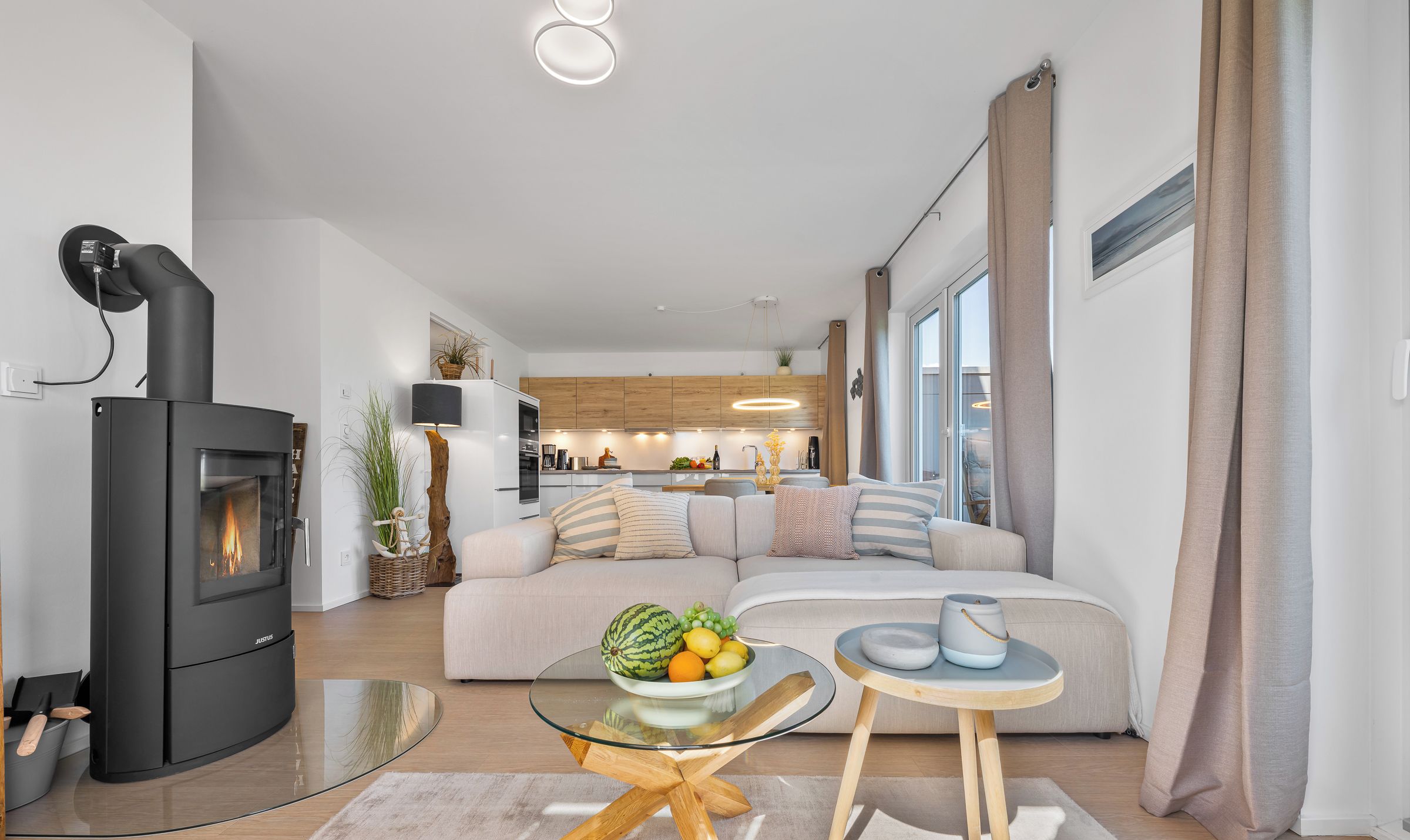 A modern living room with a black wood-burning stove, a glass coffee table, a beige sofa, and a kitchen in the background.