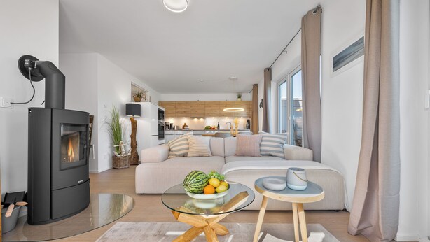 Una sala moderna con un estufa de leña negra, una mesa de centro de vidrio, un sofá beige y una cocina al fondo.