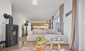 A modern living room with a black wood-burning stove, a glass coffee table, a beige sofa, and a kitchen in the background.