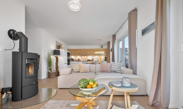 A modern living room with a black wood-burning stove, a glass coffee table, a beige sofa, and a kitchen in the background. - Ankermann II - Wood-burning stove, sauna, shower, terrace, parasol, countryside (Olpenitz)