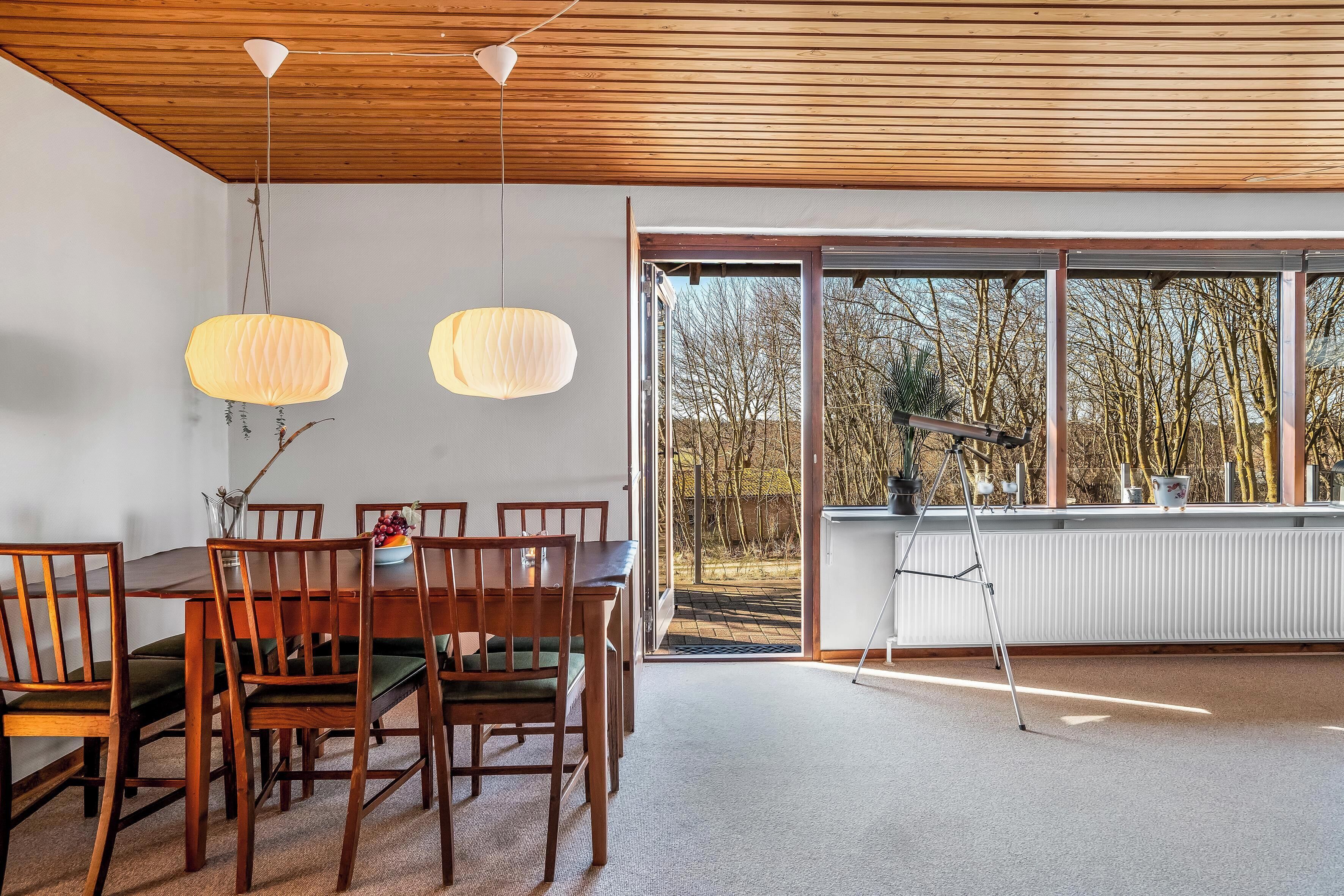 A dining area with wooden chairs and a table, large windows with a view of trees, and hanging pendant lights.