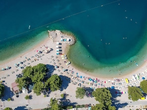 An aerial view of a beach with rows of sunbeds and umbrellas, a small rocky area, and clear blue waters.