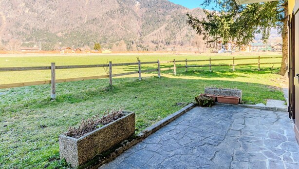 A stone patio with a view of a mountain range, a wooden fence, and a grassy field.