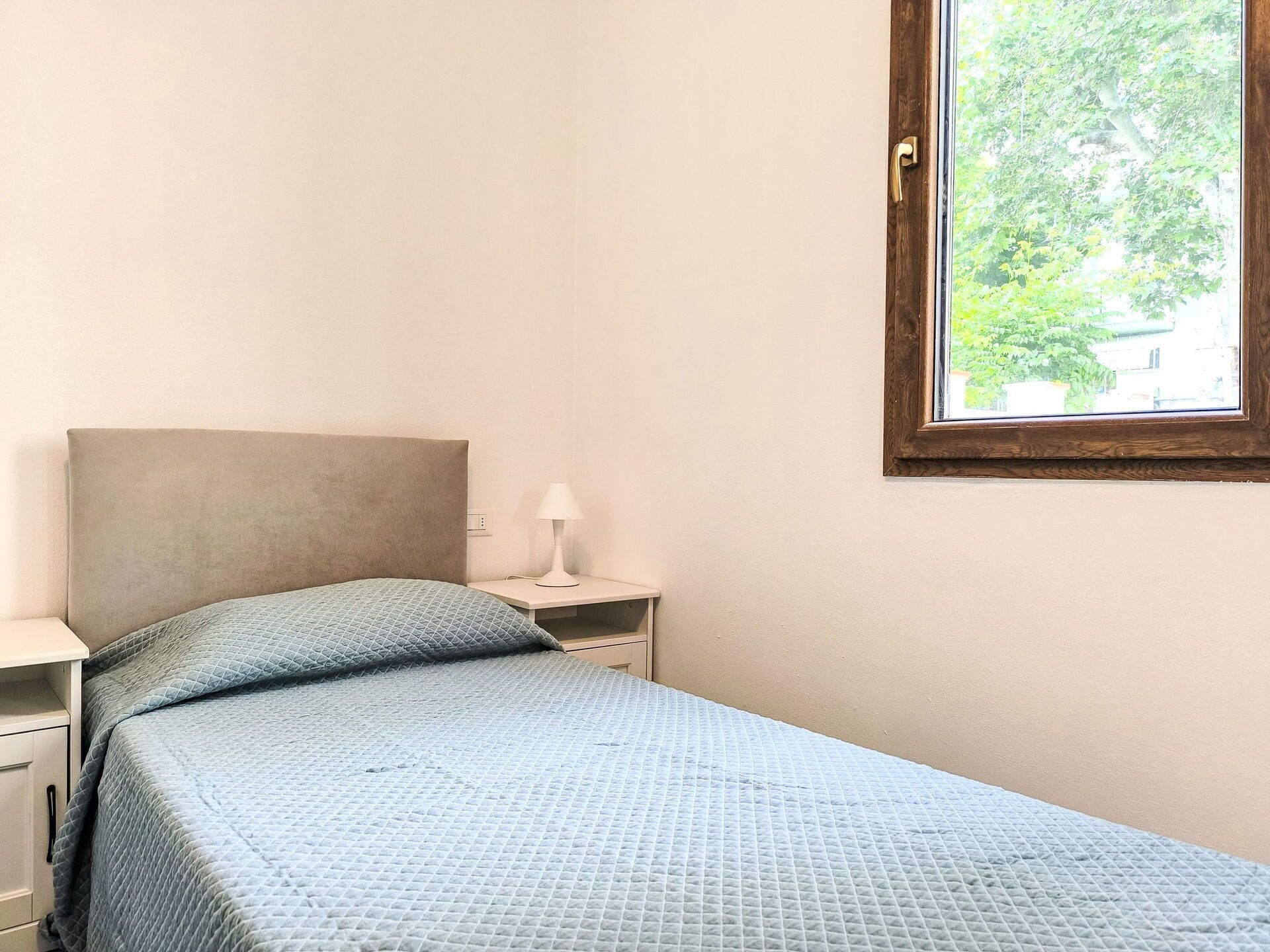 A small, neatly made bed with a blue quilt, a bedside table with a lamp, and a window with a view of trees and buildings.