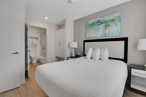 A hotel room with a large bed, a nightstand, a bedside table, a wall-mounted painting, and a bathroom visible through an open door.