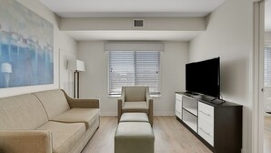 Un salon moderne avec un canapé beige, un pouf assorti, un téléviseur posé sur un meuble et un grand tableau abstrait accroché au mur.