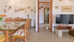 Ein modernes Wohnzimmer mit einem Glastisch, Korbstühlen, einem Fernseher und einer Küche im Hintergrund.