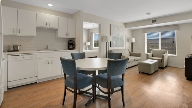A modern kitchen with white cabinets, a dining table with chairs, and a living area with a sofa and ottoman.