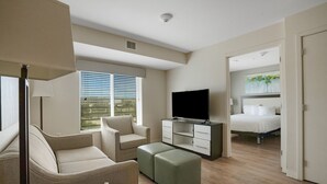 A modern hotel room with a flat-screen TV, a sofa, a small table with a telephone and a basket, and a bed with a white headboard.