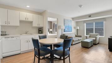 A modern kitchen with white cabinets, a dining table with four chairs, and a living area with a sofa and ottoman.
