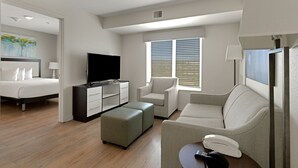 A modern hotel room with a bed, a TV, a sofa, a desk, and a window with blinds.