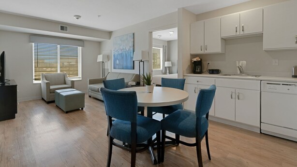 A modern kitchen with white cabinets, a dining area with a round table and blue chairs, and a living room with a sofa and a TV.