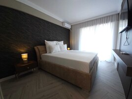 A modern hotel room with a large bed, a dark textured wall, a bedside table with a lamp, and a large window with curtains.