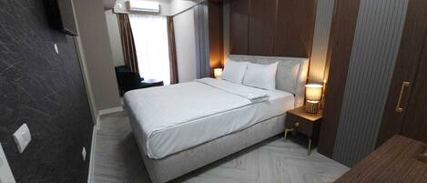 A hotel room with a large bed, a bedside table with a lamp, a TV mounted on the wall, and a window with curtains.