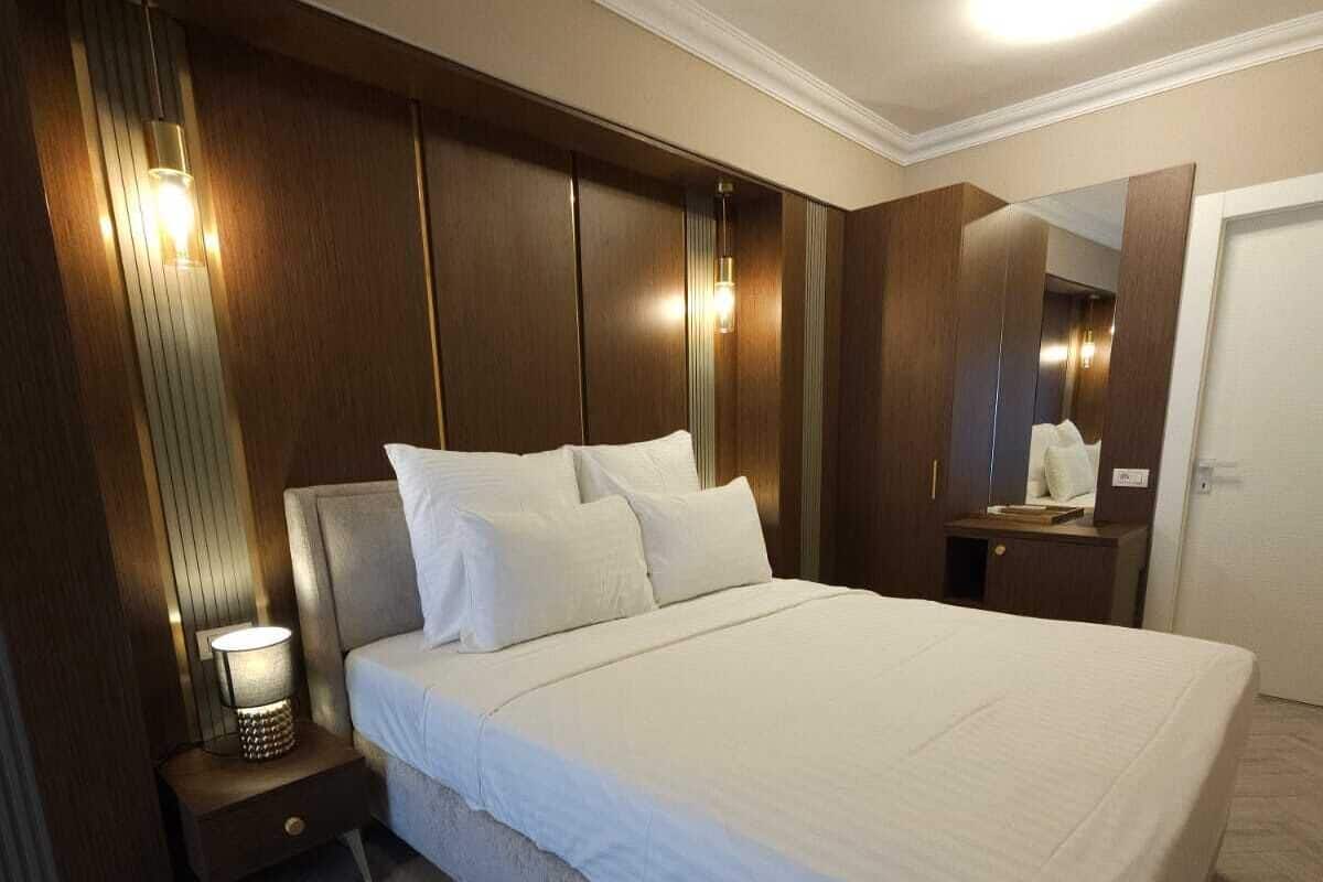 A spacious bedroom with a large bed, wooden paneling, a nightstand with a lamp, and a mirror.