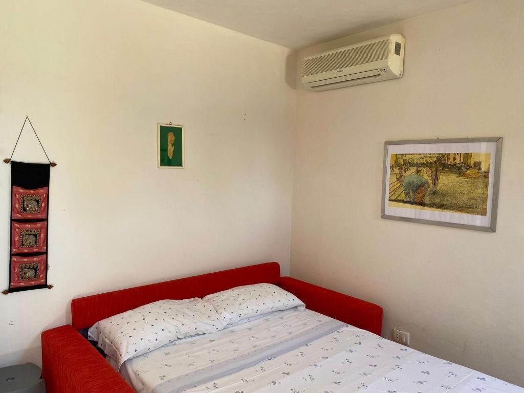 A bedroom with a red bed, a white wall, an air conditioner, a framed picture, and a green poster.