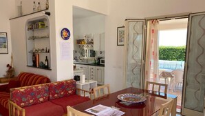 A dining area with a glass table, wooden chairs, and a red sofa. There is a kitchen area with cabinets and a microwave. A glass door leads to an outdoor area with a table and chairs. - Villa Amos Marzamemi San Lorenzo (San Lorenzo)