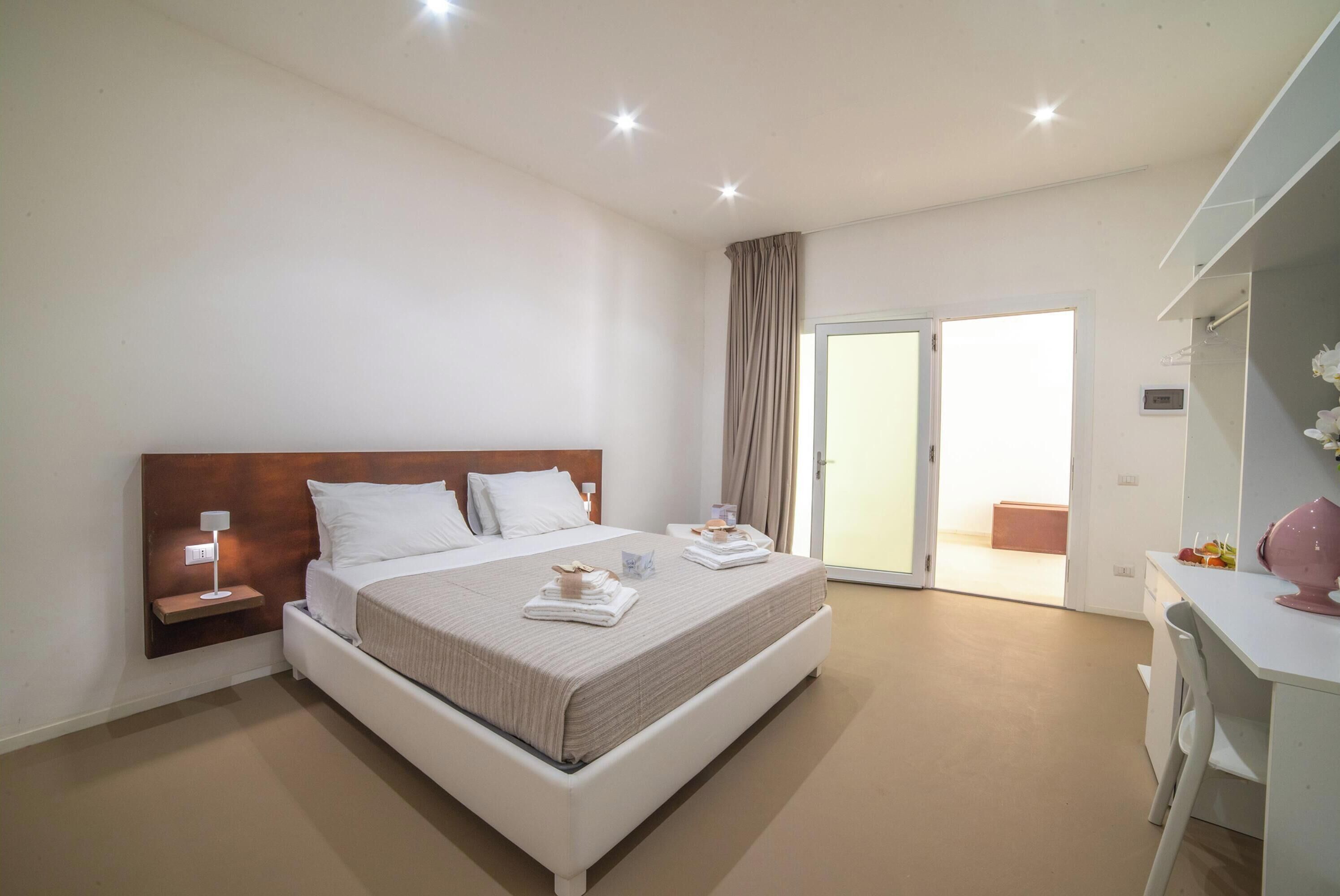 A hotel room with a large bed, a desk, a chair, and a sliding glass door leading to an outdoor area.