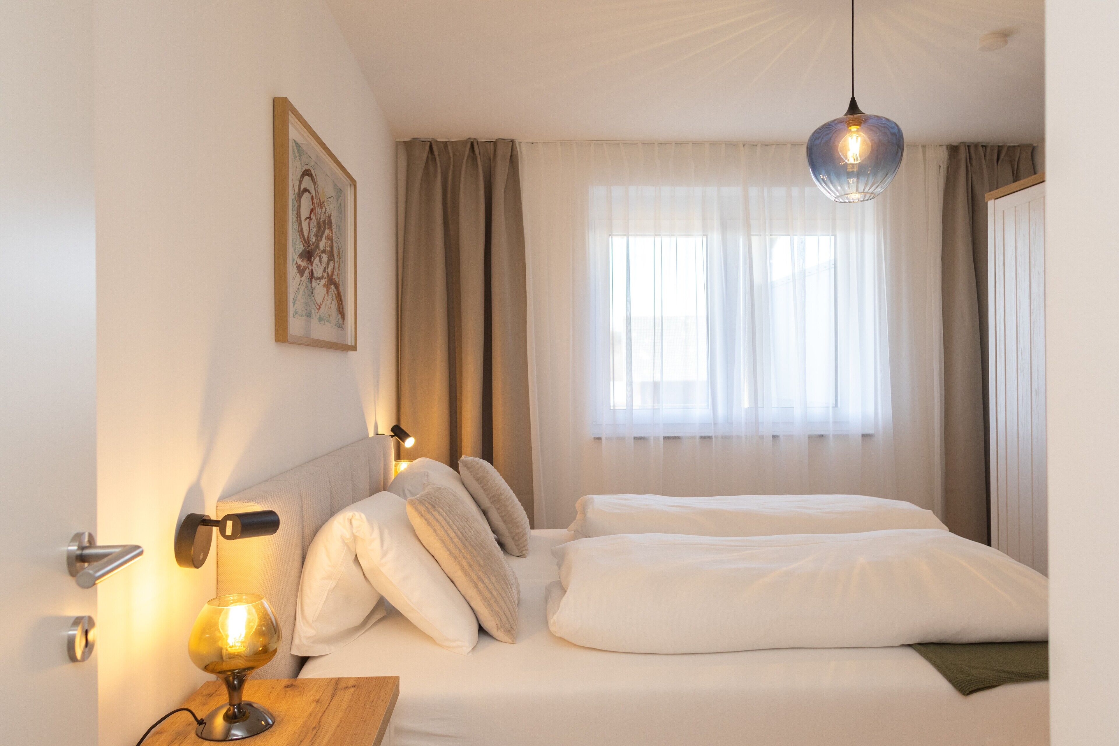 Une chambre d’hôtel avec un lit, une table de chevet avec une lampe, une fenêtre avec des rideaux et un tableau accroché au mur.