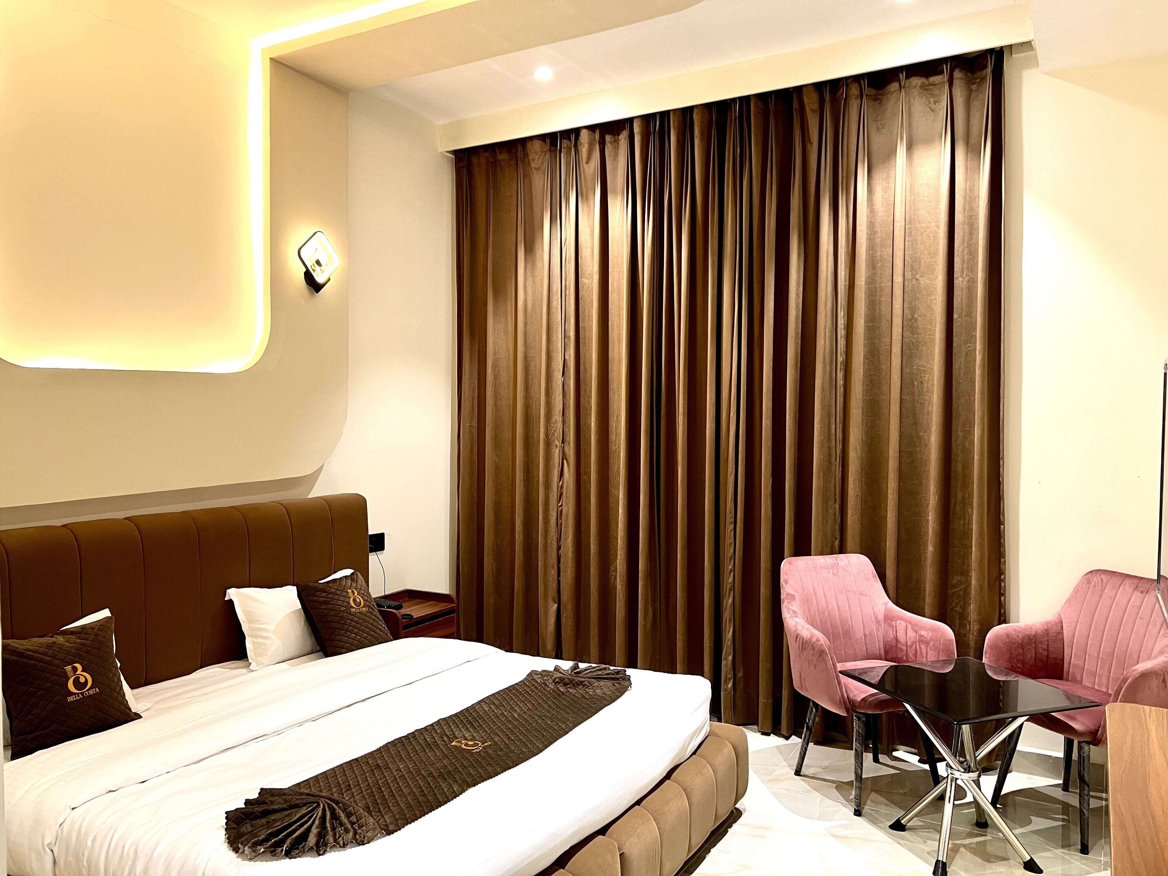A modern hotel room with a large bed, a desk, and two pink chairs.