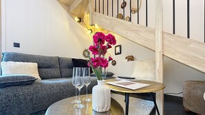 Un salon moderne avec un canapé gris, une table ronde ornée d’un vase blanc et de fleurs roses, et deux verres à vin vides.