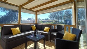 An outdoor covered patio with a wooden ceiling, a dark grey sofa with yellow cushions, a wicker chair, and a small table with wine glasses. - Comfortable mobile home near the Atlantic (Talmont-Saint-Hilaire)