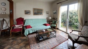 Een gezellige woonkamer met een blauwgroene bank, rode fauteuils, een salontafel en een vloerkleed met een patroon. Er hangen afbeeldingen in lijsten aan de muur, een lamp en een raam met gordijnen.